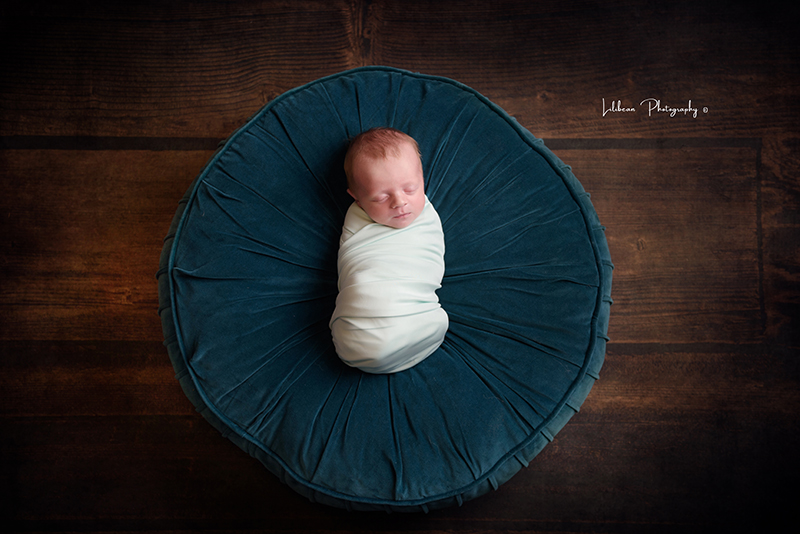 Newborn baby swaddled on top of a round blue pillow on top of wood floor
